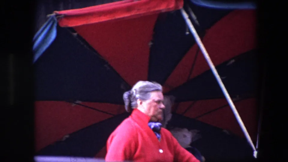 The final 1967 Brussels frame shows the older woman in profile under the large red and dark umbrella, with her gray hair tied back and the urban background mostly in shadow.