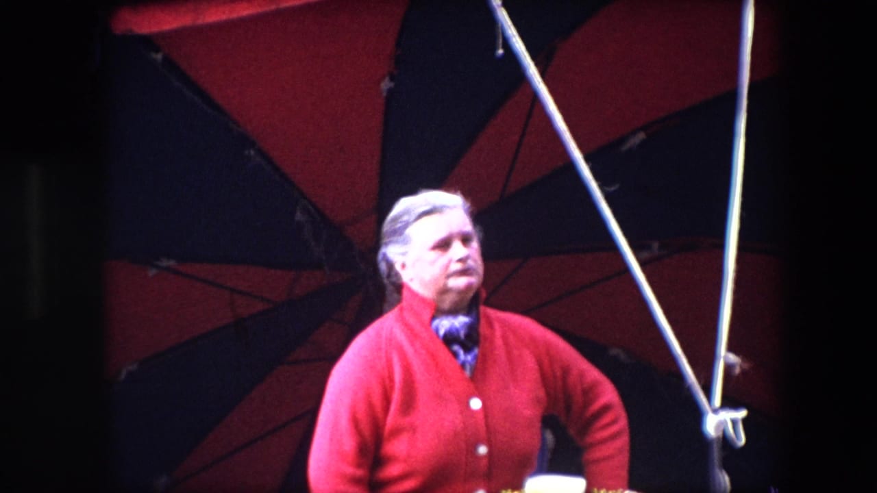 In Brussels home-movie footage from 1967, an older woman in a red cardigan and patterned scarf stands under a red and black striped canopy with tent poles around her.