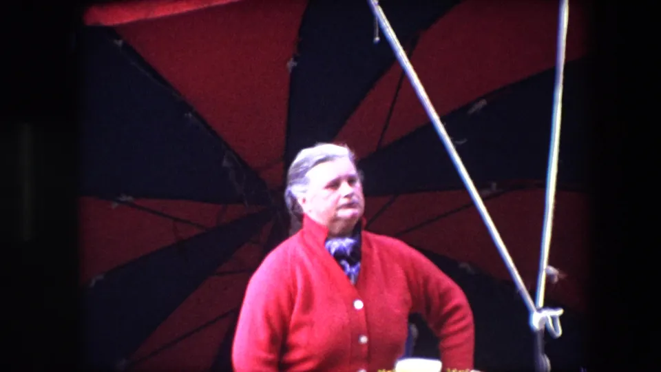 In Brussels home-movie footage from 1967, an older woman in a red cardigan and patterned scarf stands under a red and black striped canopy with tent poles around her.