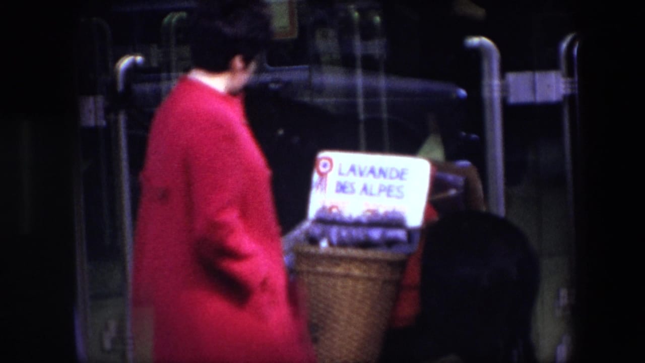 In 1967 Brussels footage, a traveler in a red coat stands near glass vehicle doors with a wicker basket labeled LAVANDE DES ALPES and lavender bundles.
