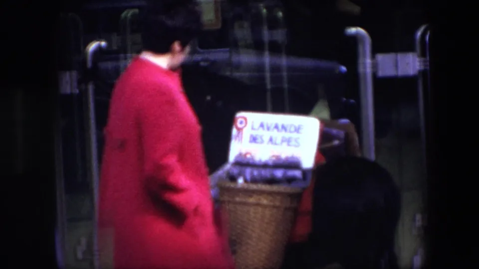 In 1967 Brussels footage, a traveler in a red coat stands near glass vehicle doors with a wicker basket labeled LAVANDE DES ALPES and lavender bundles.