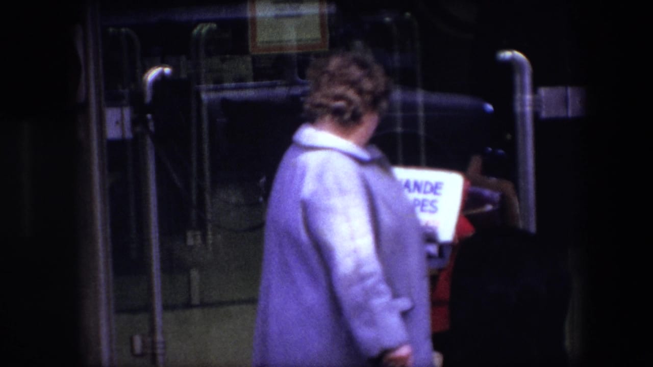A woman in a pale coat moves past the LAVANDE DES ALPES basket at a glass vehicle entrance in the Brussels 1967 home-movie scene.