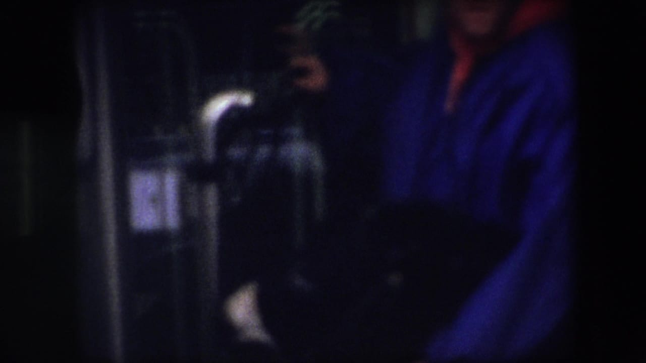 In 1967 Brussels home-movie footage, a person in a bright blue jacket and red hood or scarf stands close to dark interior fixtures in a soft, underexposed frame.