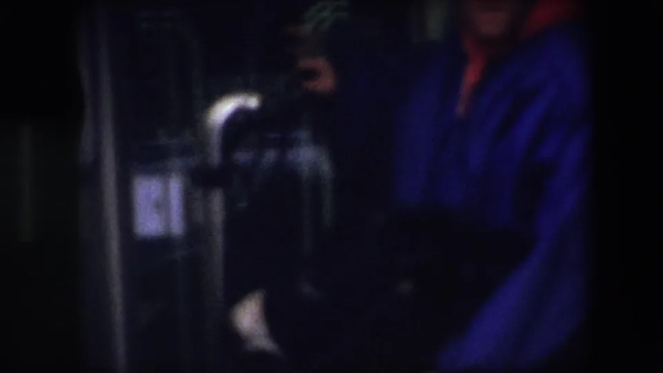 In 1967 Brussels home-movie footage, a person in a bright blue jacket and red hood or scarf stands close to dark interior fixtures in a soft, underexposed frame.