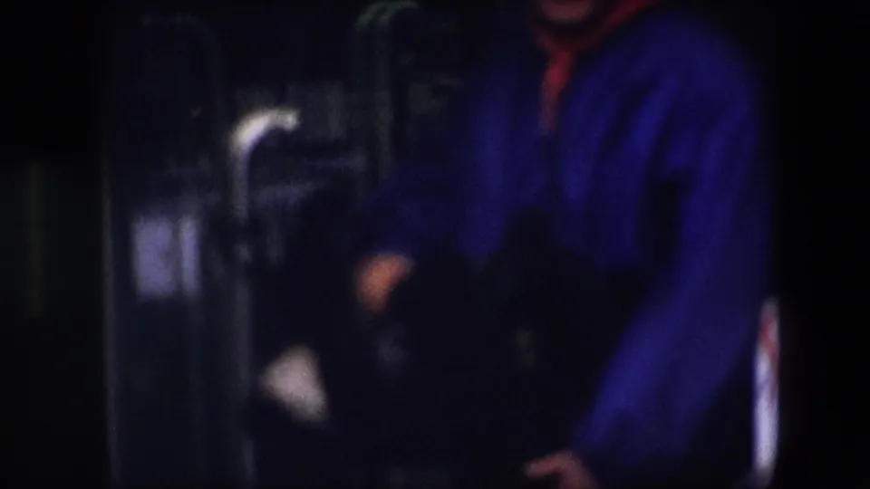 A cropped figure in a bright blue jacket and red hood or scarf appears to move or gesture inside a shadowed Brussels interior captured on 8mm film.