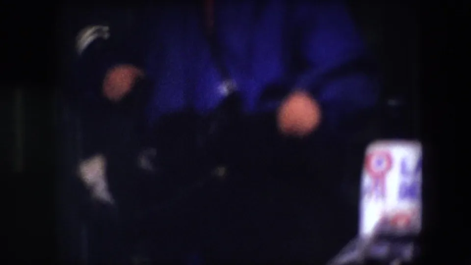 The camera frames the blue-clad person very closely, with hands, dark clothing, and a small partially readable sign or label visible near the lower right.