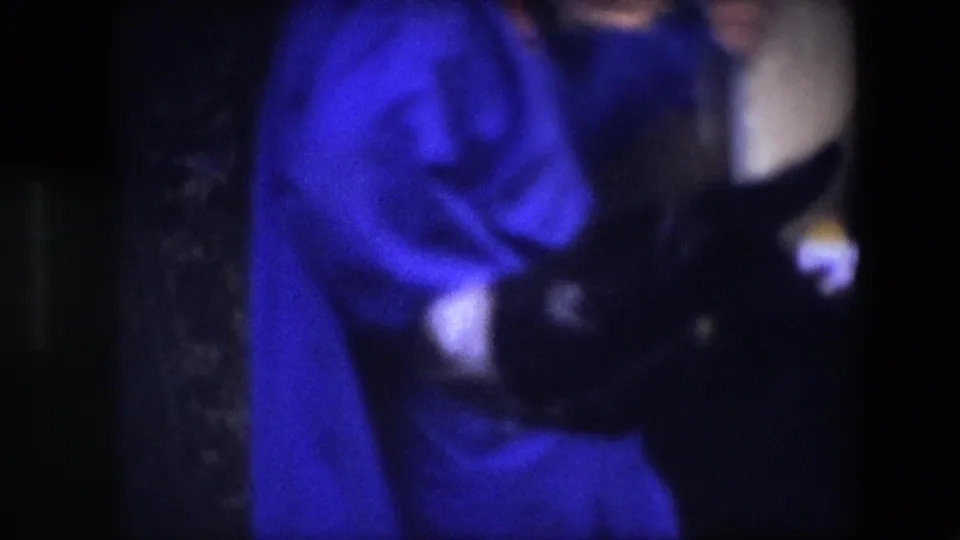 This Brussels 1967 home-movie frame crops the figures tightly, showing the woman’s bright blue dress and the neighboring person’s dark jacket in soft, grainy low light.