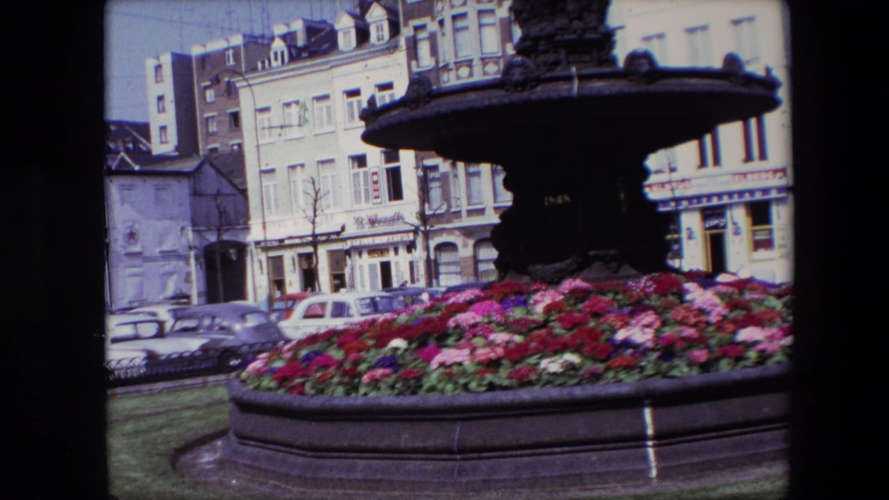 1967 Brussels 8mm frame showing an ornate dark fountain above red, pink, and purple flowers, with parked cars and storefront buildings in the background.