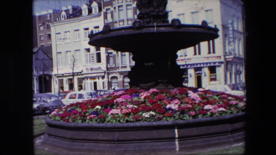 1967 Brussels 8mm frame centered on the fountain basin and bright flower bed, with classic cars and shopfront facades visible across the street.