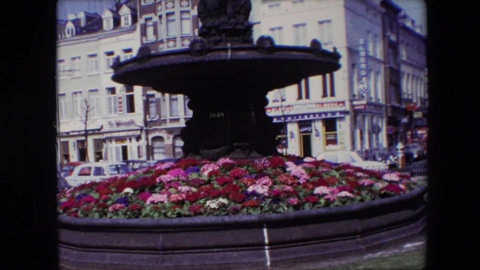 1967 Brussels 8mm frame with the camera closer to the fountain, showing dense flowers in the foreground and masonry buildings with signs behind.