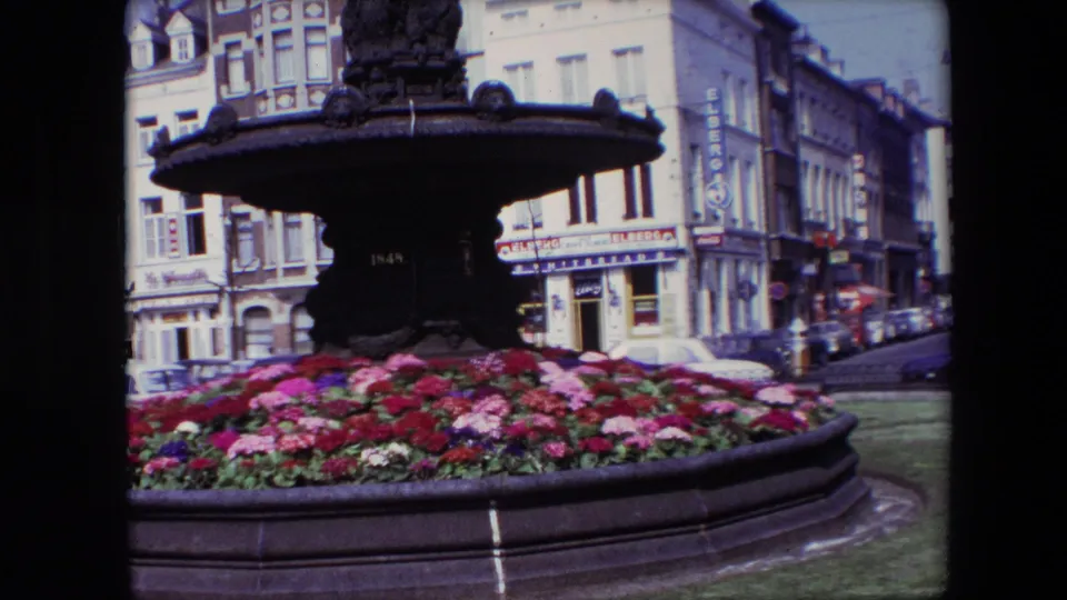 1967 Brussels 8mm frame showing the fountain and flower bed from a slightly shifted angle, with parked cars, storefront signs, and city buildings continuing down the street.