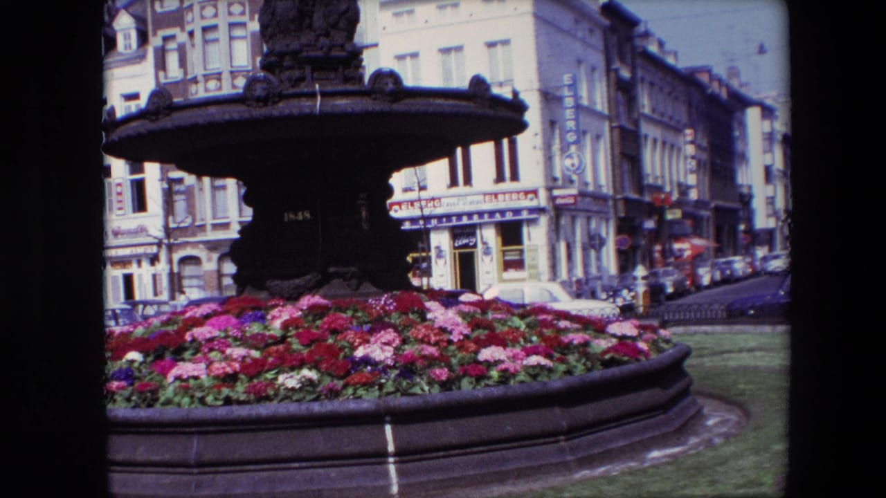 1967 Brussels home-movie frame showing an ornate dark fountain surrounded by bright flower beds, with shopfronts, parked cars, and European facades behind it.