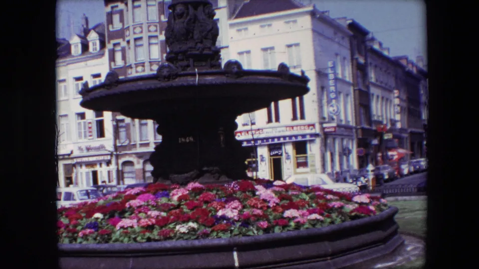 1967 Brussels frame with the fountain centered above colorful flower beds, backed by commercial buildings, visible signage, and parked street cars.
