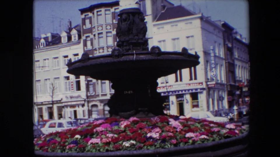1967 Brussels home-movie view looking across vivid flowers toward an ornate fountain, with parked cars and historic street buildings visible in the background.