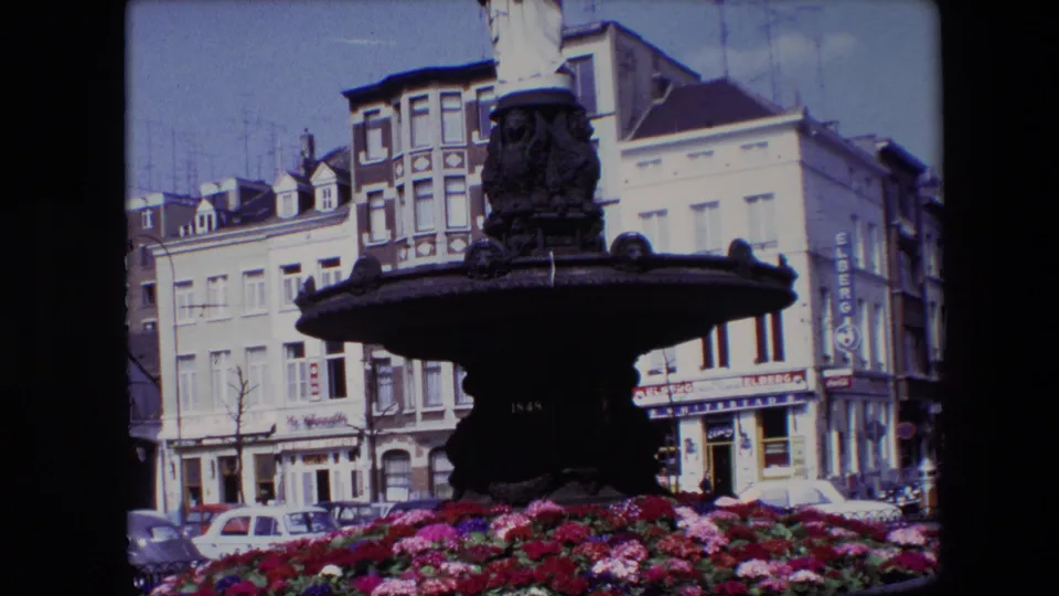 Final 1967 Brussels frame showing the fountain and surrounding flower bed from a steady visitor viewpoint, with facades, signage, and parked cars behind it.