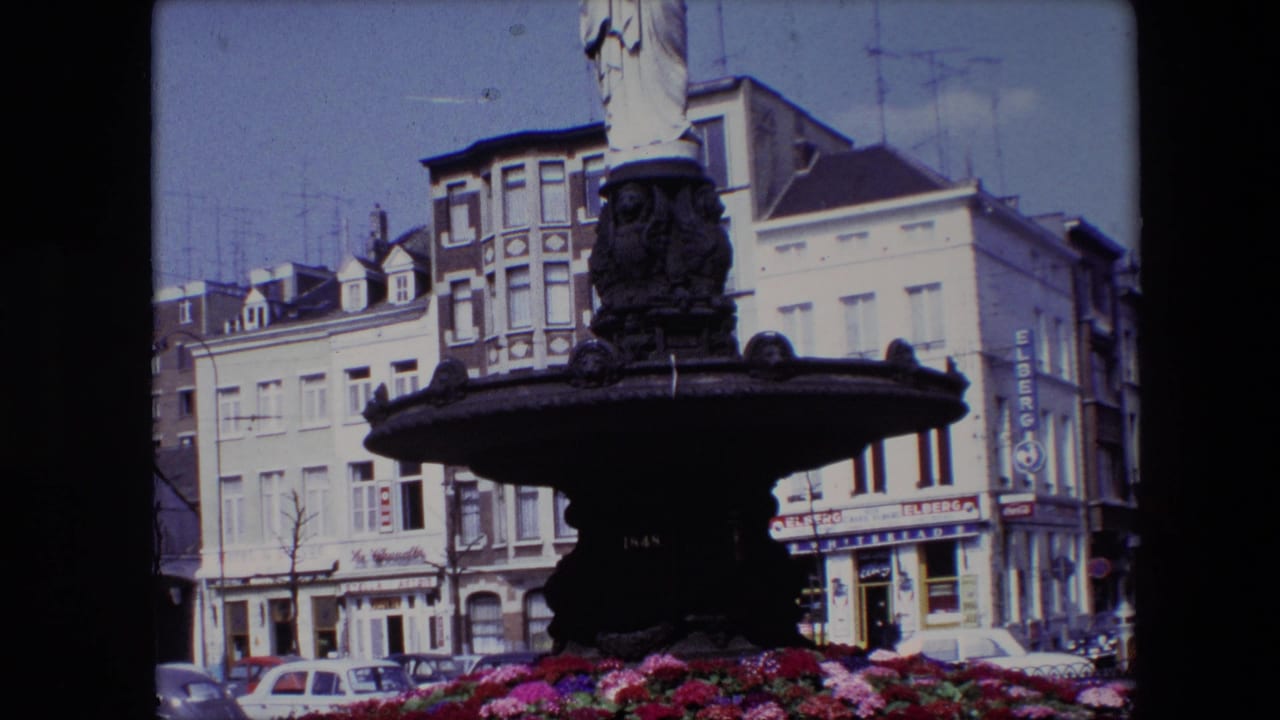 1967 Brussels 8mm frame showing a dark ornate fountain topped by a classical robed female statue, with red and pink flowers, parked cars, and multi-story buildings behind it.