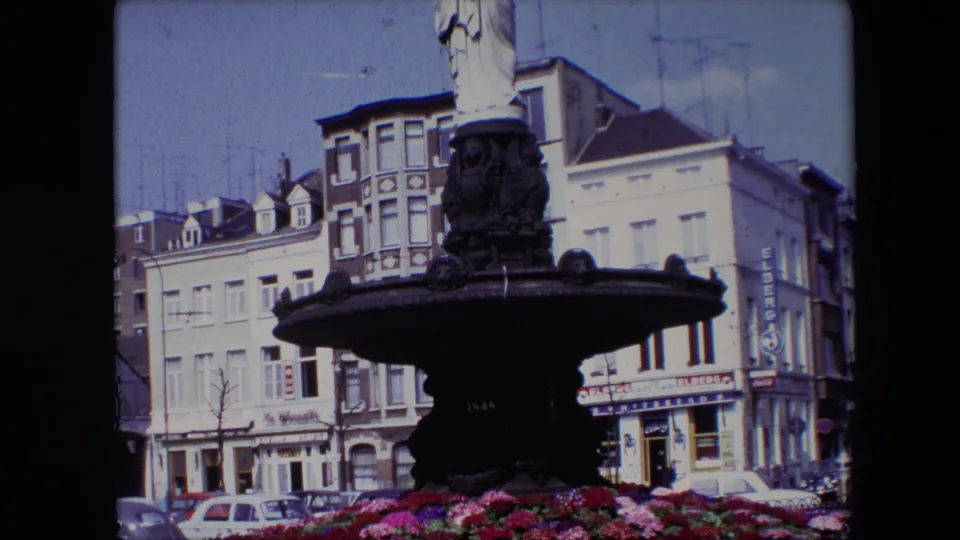 1967 Brussels 8mm frame showing a dark ornate fountain topped by a classical robed female statue, with red and pink flowers, parked cars, and multi-story buildings behind it.