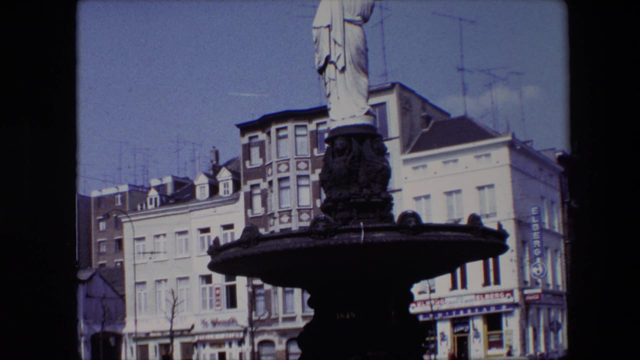 1967 Brussels 8mm frame with the fountain basin and robed statue partly cropped, set before urban facades, parked cars, rooftop antennas, and visible shop signs.