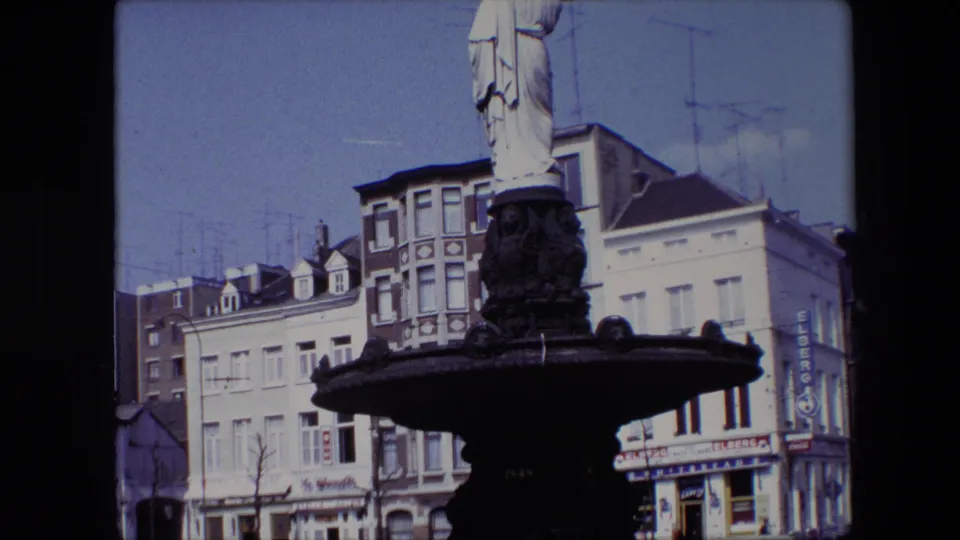 1967 Brussels 8mm frame with the fountain basin and robed statue partly cropped, set before urban facades, parked cars, rooftop antennas, and visible shop signs.