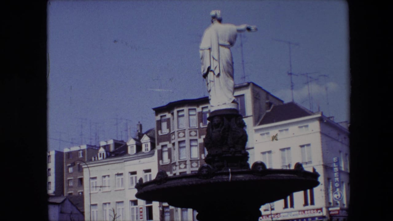Fuller view of robed statue on fountain with buildings and antennas behind.