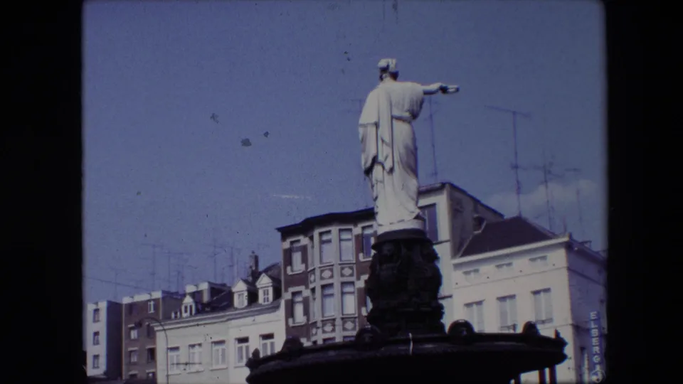 1967 Brussels 8mm frame with the statue and dark fountain basin seen from below, backed by blue sky, rooftop antennas, multi-story buildings, and visible commercial signage.