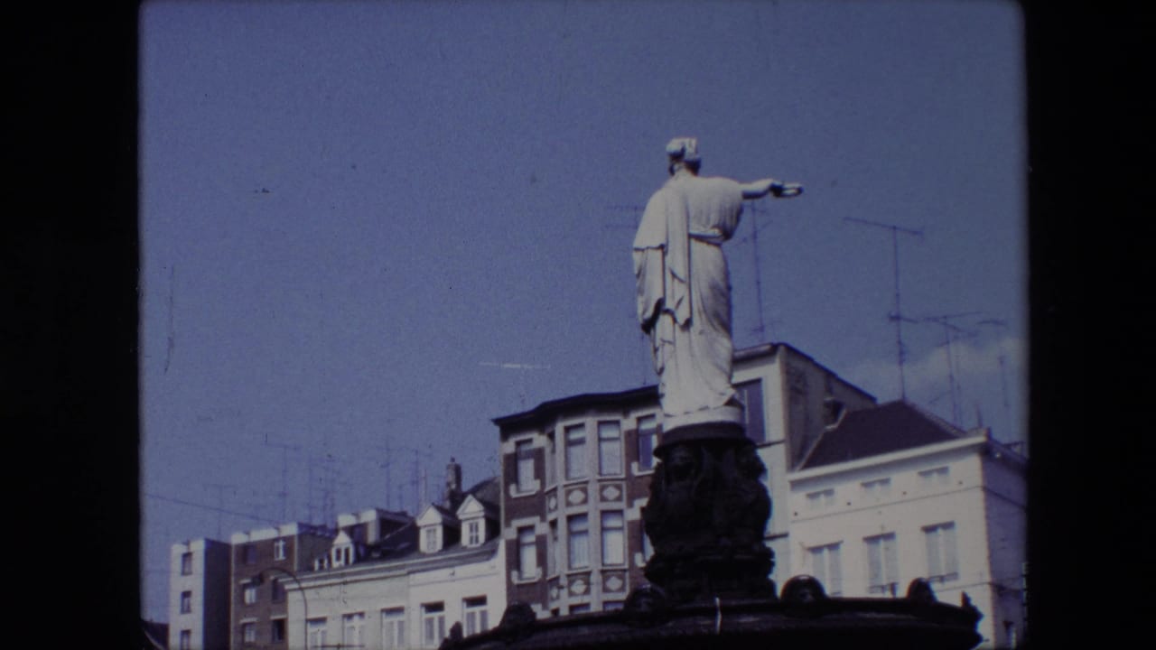 1967 Brussels home-movie frame showing a white stone statue on a fountain base, with multi-story buildings and rooftop antennas behind it.