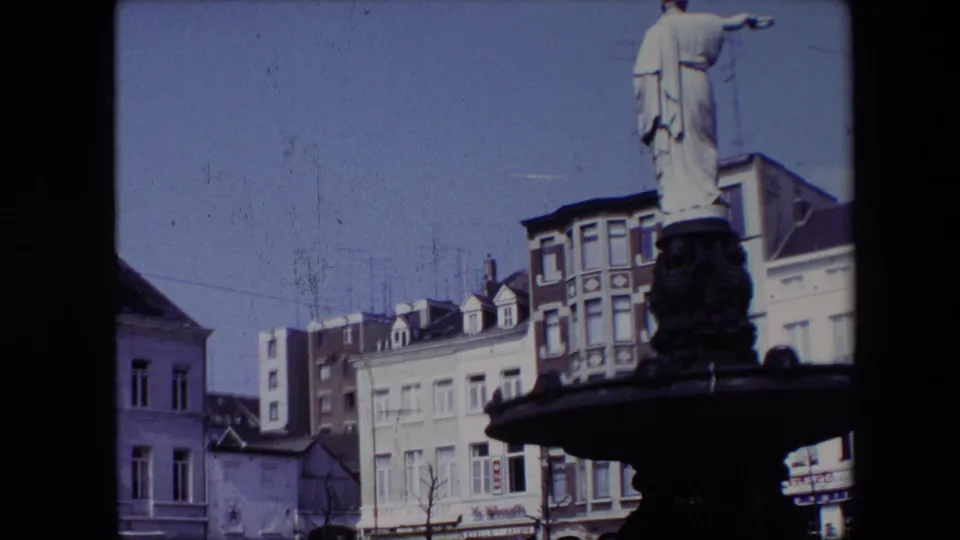 1967 Brussels home-movie frame during the pan, showing the fountain statue, nearby facades, rooftop antennas, and part of a street-level building edge.