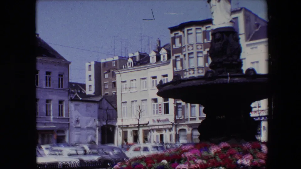 1967 Brussels frame showing the fountain statue at the right edge, city buildings behind it, parked vintage cars, bare trees, and a bright flower bed below.