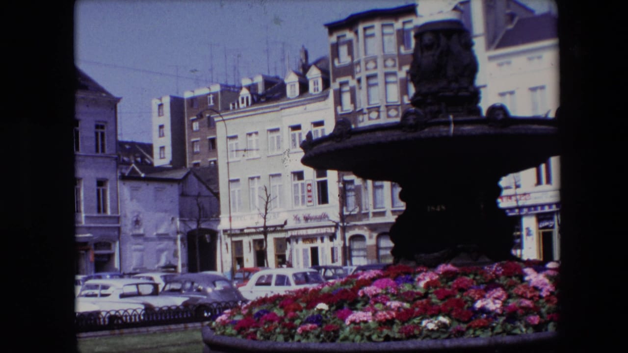 1967 Brussels 8mm frame showing an ornate dark fountain, colorful flower bed, parked cars, and multi-story street buildings in the background.