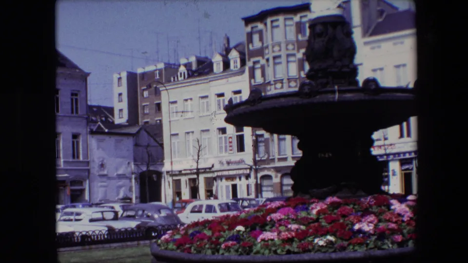 1967 Brussels 8mm frame showing an ornate dark fountain, colorful flower bed, parked cars, and multi-story street buildings in the background.