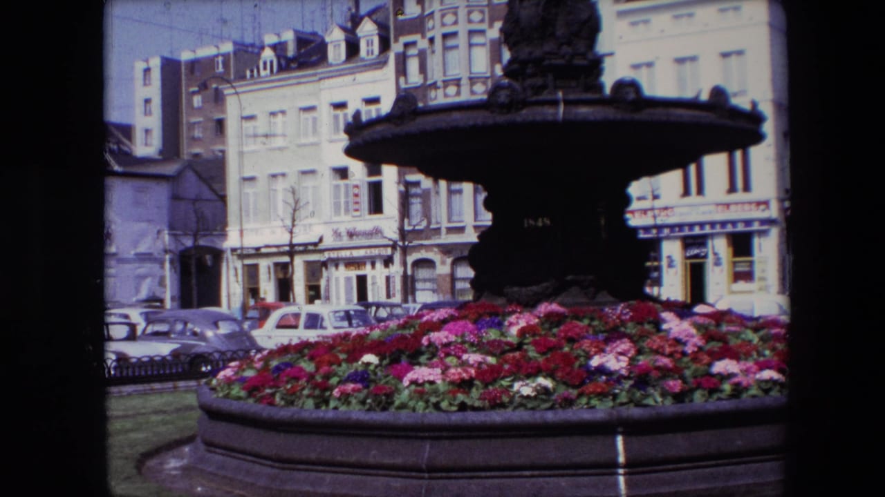 1967 Brussels 8mm frame with the fountain basin dominating the foreground, bright flowers below, and parked cars and shopfront buildings behind.