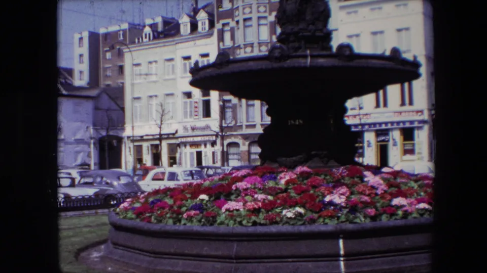1967 Brussels 8mm frame with the fountain basin dominating the foreground, bright flowers below, and parked cars and shopfront buildings behind.