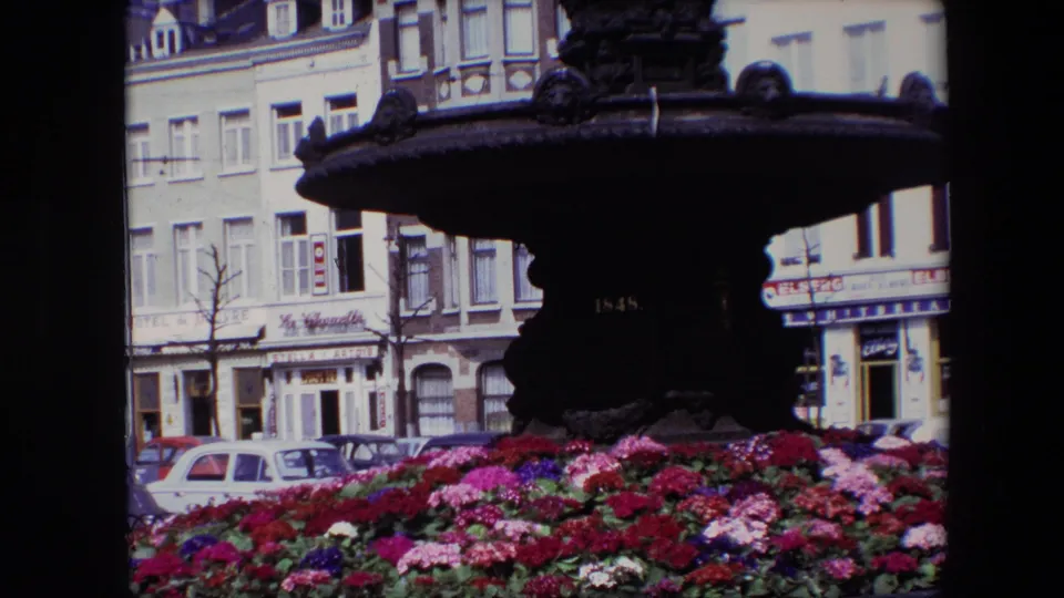 1967 Brussels 8mm frame showing a close view of the fountain base and basin above vivid flowers, with parked cars and street-facing buildings in the background.