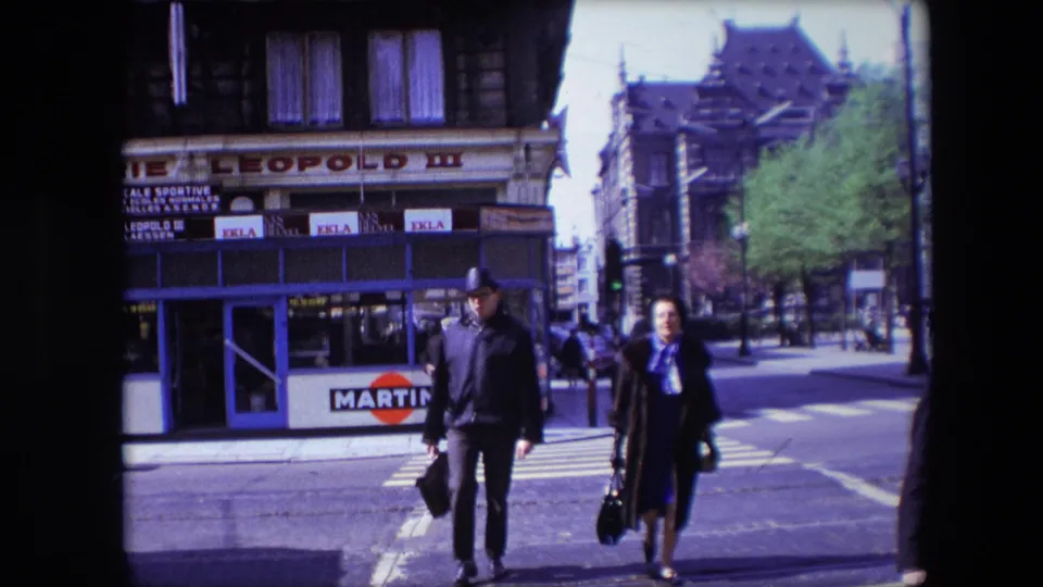 In the 1967 Calais street scene, a man in a hat and a woman in a dark coat approach the camera across a zebra crossing with shopfronts behind them.