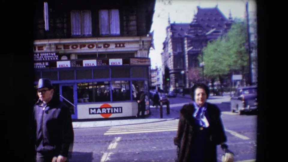 1967 Calais street corner with a woman in a dark coat and purple scarf crossing near Brasserie Leopold III as a man in a fedora passes in front of Martini advertising.