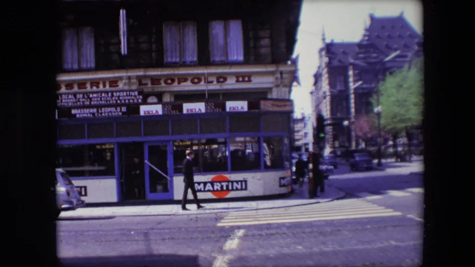 Street-level 1967 Calais view of a man walking across the zebra crossing in front of the blue Brasserie Leopold III facade, with a small car and Martini sign visible.