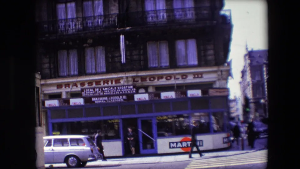 Wider slightly tilted home-movie frame of Brasserie Leopold III in Calais with Martini advertising, a light-colored car, pedestrians on the crossing, and surrounding older buildings.