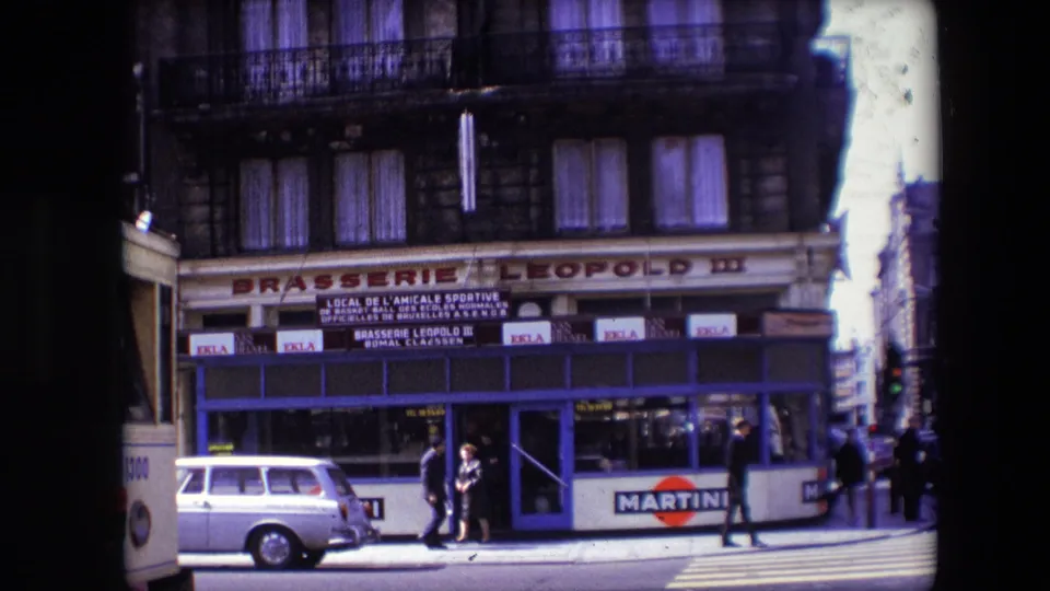 1967 Calais street view showing Brasserie Leopold III with blue storefront trim, pedestrians on the sidewalk, a light station wagon, and Martini advertising along the lower facade.