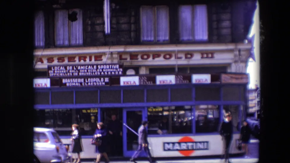 1967 Calais sidewalk scene outside Brasserie Leopold III, with several pedestrians passing the blue-trimmed frontage and Martini branding visible on panels below the windows.