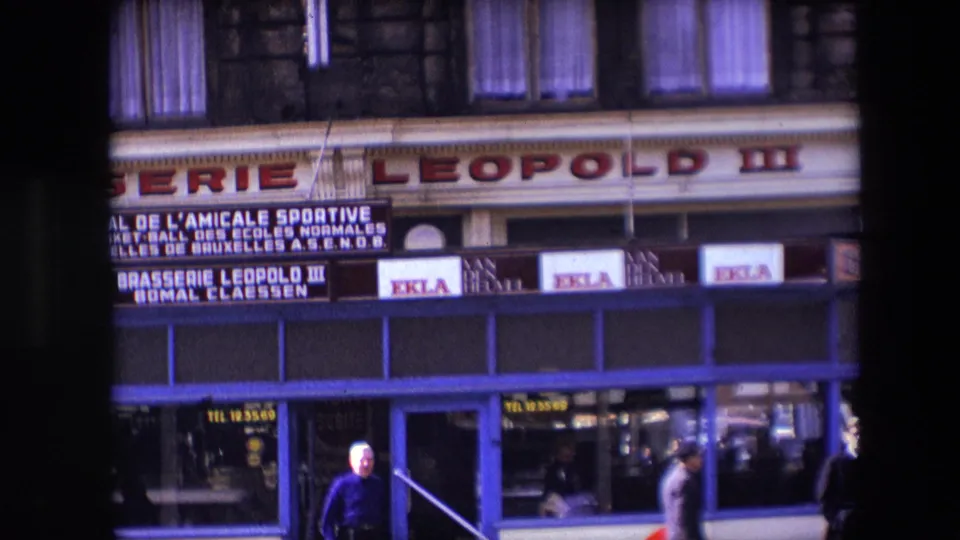 Closer 1967 Calais view of Brasserie Leopold III signage and blue storefront windows, with pedestrians at the edge of the frame and cafe-related lettering visible near the entrance.