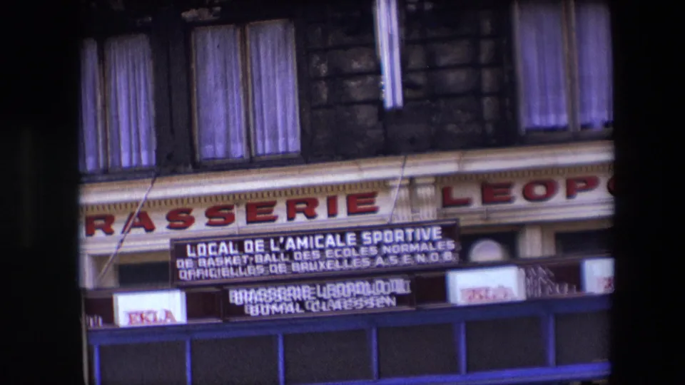 Tight 1967 Calais frame on the Brasserie Leopold III facade, showing painted sign lettering, upper windows with curtains, blue storefront trim, and visible advertising panels.
