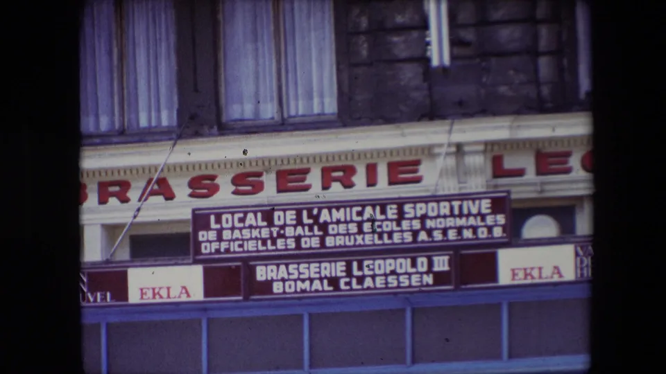 1967 Calais storefront detail showing large brasserie lettering, the French A.S.E.N.O.B. association notice, EKLA advertising panels, blue shopfront framing, and curtained upper windows.