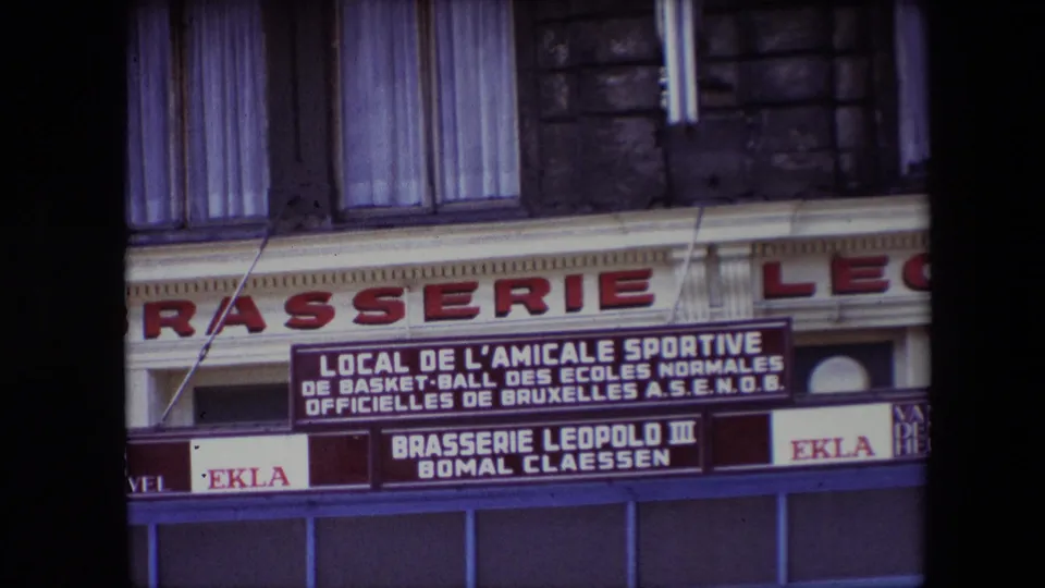 1967 8mm travel frame showing a European brasserie exterior with a French-language sign for a Brussels school basketball sports association and Ekla beer branding.