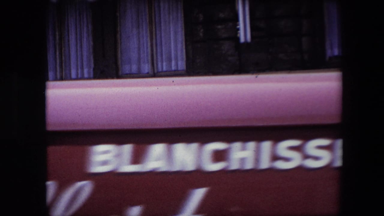 Soft 8mm frame from the same travel clip with a blurred Blanchisserie storefront sign, pink awning, upper windows, and heavy motion blur.