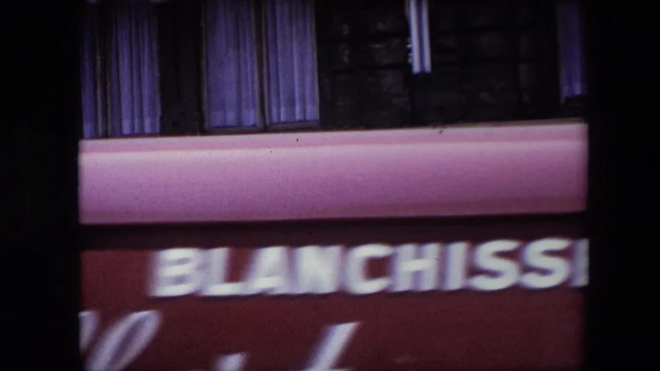 Soft 8mm frame from the same travel clip with a blurred Blanchisserie storefront sign, pink awning, upper windows, and heavy motion blur.