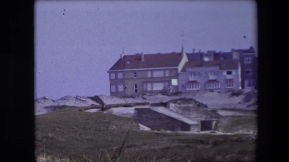 1967 8mm coastal view, reportedly from the Calais travel footage, with European-style buildings behind dunes, grass, and low concrete bunker-like forms.