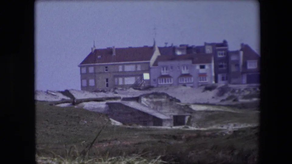 Late frame of the 1967 8mm clip showing a seaside row of buildings beyond grass, sand dunes, and low concrete bunker-like structures.