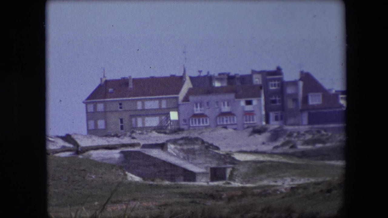 1967 Calais home-movie frame showing a large coastal building with red tile roofs beyond sandy dunes and a low concrete structure.