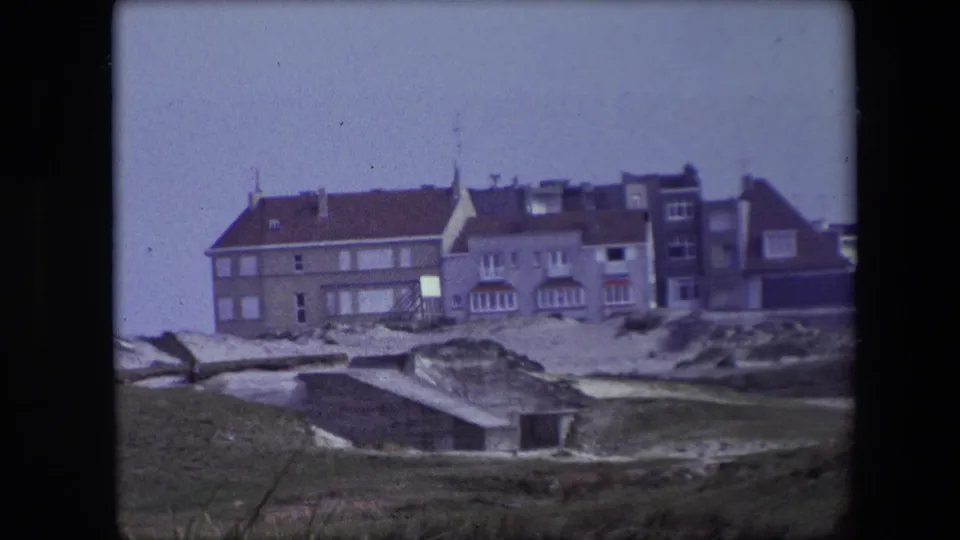 1967 Calais home-movie frame showing a large coastal building with red tile roofs beyond sandy dunes and a low concrete structure.