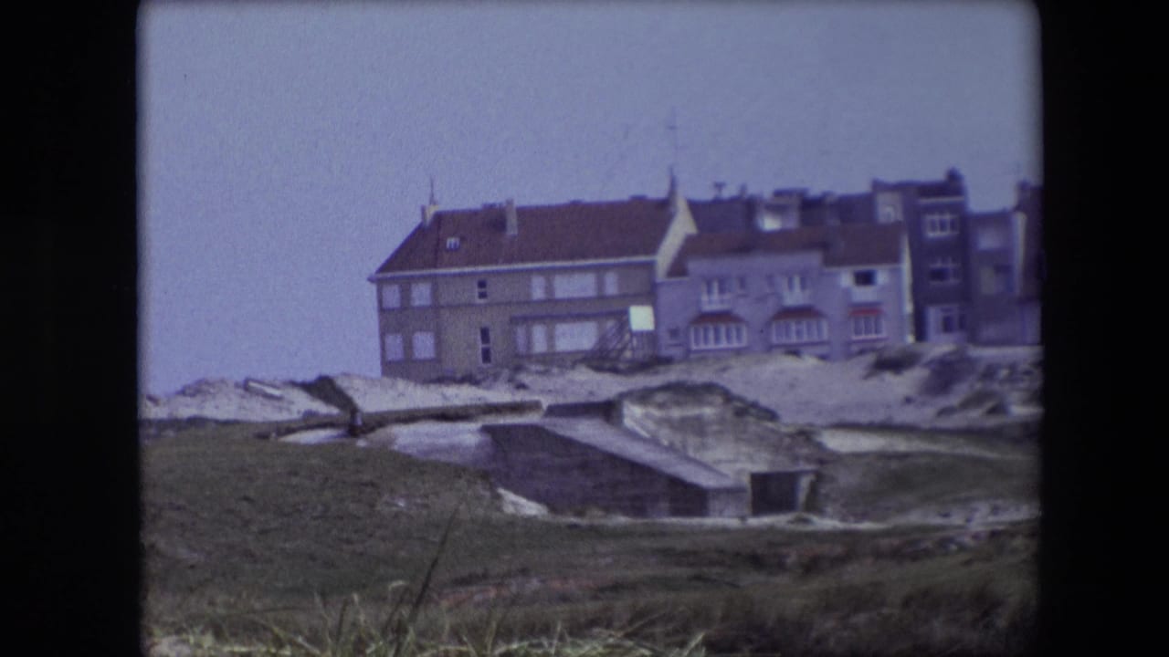 Coastal building with rooflines and dunes viewed from a grassy foreground.